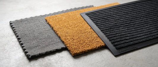 Drei verschiedene langlebige Fußmatten im Vergleich auf hellem Betonboden: Eine graue Stoff-Schmutzfangmatte, eine braune Kokosmatte und eine schwarze, gerippte Bodenständig Schmutzfangmatte mit Gummirand.