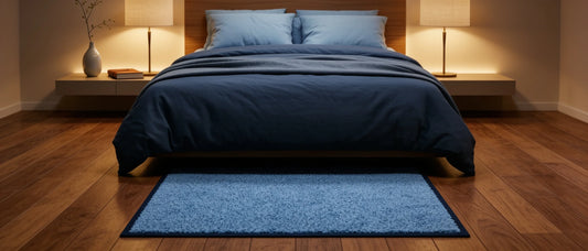 Hellblauer Schlafzimmerteppich auf dunklem Parkettboden vor einem modern gemachten Bett mit blauer Bettwäsche und gemütlicher Nachttischbeleuchtung.