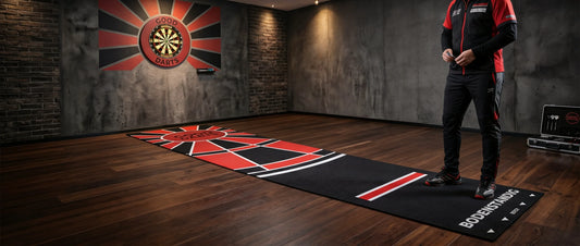 Professionelle Bodenständig Darts-Teppichmatte in Schwarz und Rot mit Abwurflinie (Oche) auf Holzboden, im Hintergrund eine Dartscheibe an einer Klinkerwand und ein Dartspieler.
