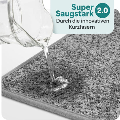 Wasser wird auf eine hellgraue Badematte gegossen und sofort absorbiert, dank innovativer Kurzfasern mit starker Saugkraft fuer das Badezimmer