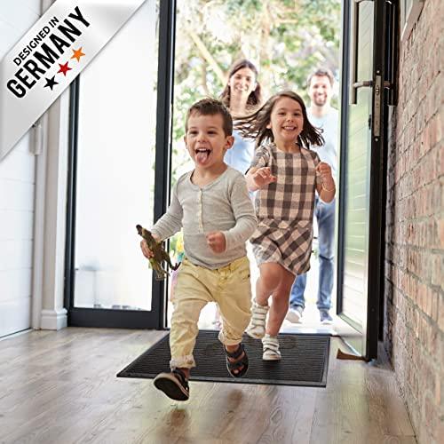 Kinder rennen in den Flur und laufen ueber eine schwarze Fussmatte, waehrend Erwachsene an der offenen Haustuer stehen.
