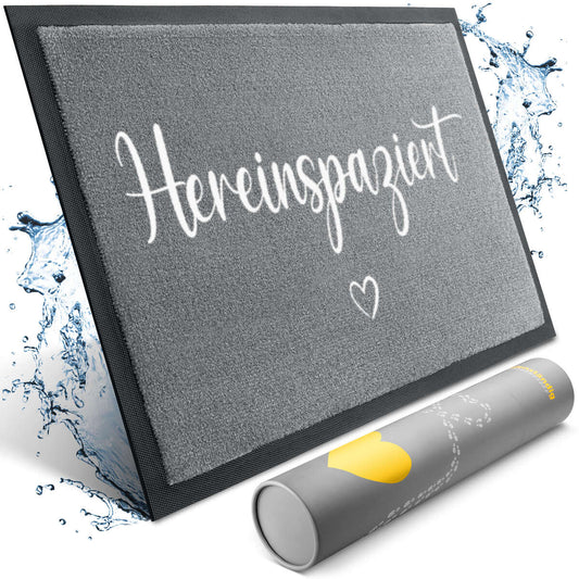 Schräge Perspektive einer Fußmatte mit dem Schriftzug Hereinspaziert. Im Hintergrund spritzt Wasser und im Vordergrund liegt die Verpackung.