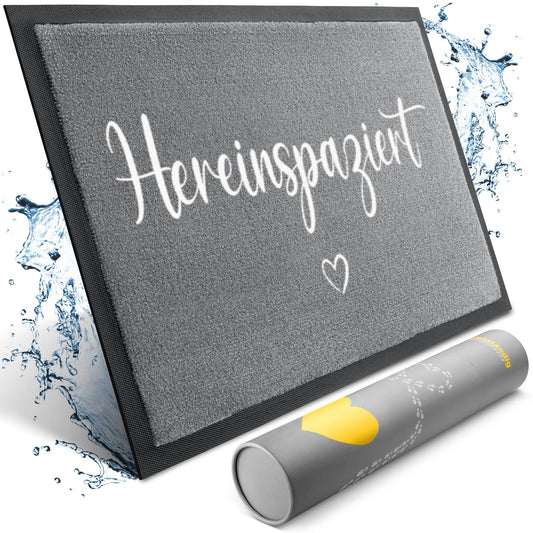 Graue Fußmatte mit weißem Schriftzug „Hereinspaziert“ und Herz, umgeben von Wasserspritzern, davor eine Versandrolle