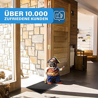 Hund im Kostuem sitzt auf einer schwarzen Fussmatte im Hauseingang, die rutschfest ist und Schmutz aufnimmt.
