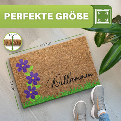 Kokosmatte „Willkommen“ mit lila Blumen und Gras, Maße 40 x 60 cm und 1,5 cm dick, daneben Schuhe und Pflanze
