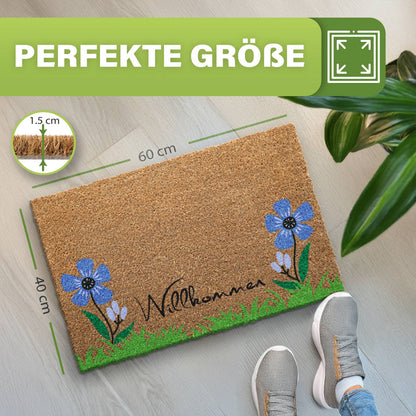 Kokosmatte „Willkommen“ mit blauen Blumen und Gras, Maße 40 x 60 cm, 1,5 cm dick, neben Pflanze und Schuhen