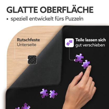 Schwarze Puzzlematte mit glatter Oberfläche, rutschfester Unterseite auf Holztisch und Hand, die lila Puzzleteile verschiebt